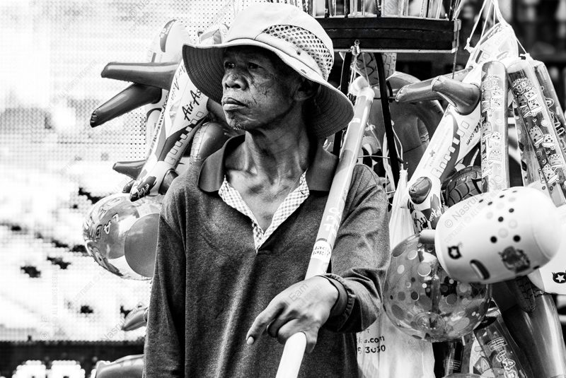 Vendor Amidst Inflatable Toys Vendor Amidst Inflatable Toys - Fine Art Photography Print, Limited Edition Photography, Documentary Photography, Black and White Photography, Art Photography for Sale