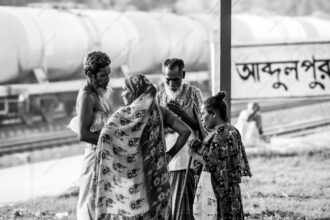 Four Figures Beside the Abdulpur Sign - Four Figures Beside the Abdulpur Sign, Nasrul Eam photography, limited edition fine art print, monochrome visual storytelling, lifestyle art photography, Bangladesh train station print